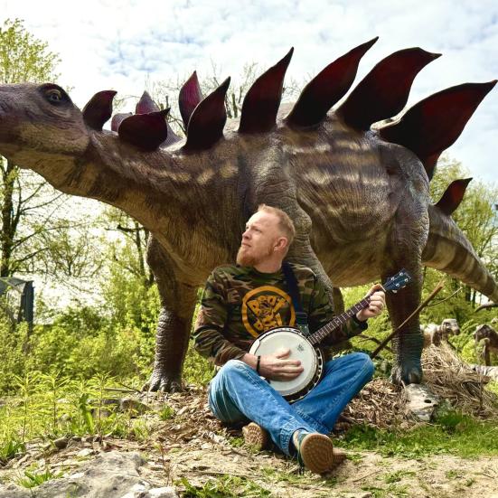 Foto af mand med guitar der sidder foran en udstoppet Stegosaurus. 