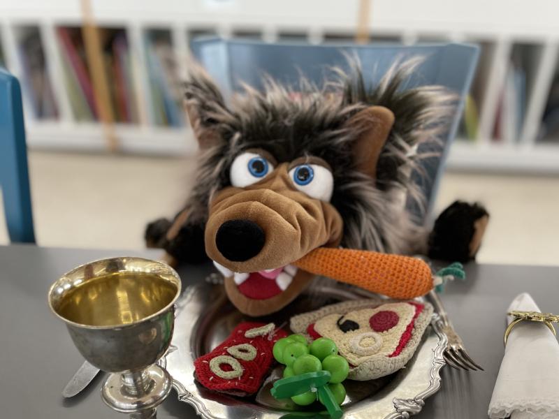 Ulven, som er et tøjdyr, sidder til bords med en gulerod i flaben.