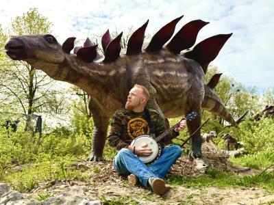 Foto af mand med guitar der sidder foran en udstoppet Stegosaurus. 
