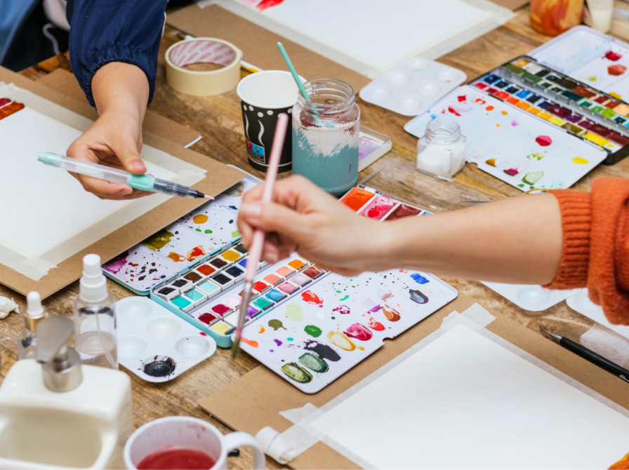 Foto af bord med akvarelmalerier, hænder der holder pensler