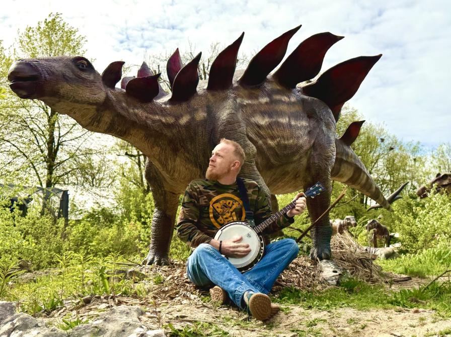 Foto af mand med guitar der sidder foran en udstoppet Stegosaurus. 